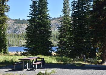 A shaded campsite with a picnic table and a beautiful mountain lake in the background.