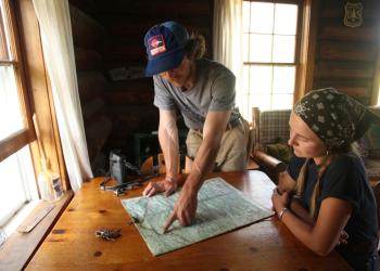 Two people sit inside a cabin and look at a map spread out on a table.