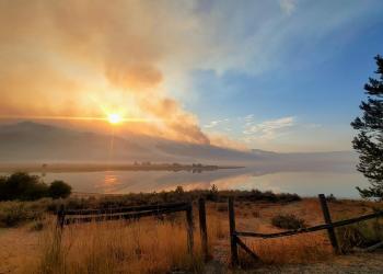 Wildfire smoke penetrates the rising sun and is reflected in a lake at the base of the mountain.
