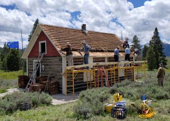 A crew of people stand on scaffolding along the side of a historic cabin and work to replace the building's roof. Grass and sage surround the facility, and a number of tools sit on the grass. In the background, conifer trees in a variety of sizes appear before a mostly cloud-filled sky. .
