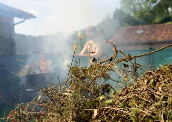 A pile of lawn debris sits in the foreground. In the back, a man stands near a burn barrel with an active fire behind a cloud of smoke. 