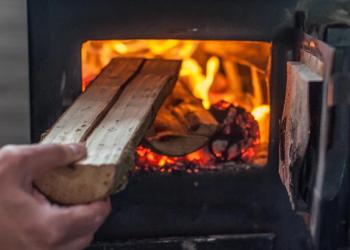 A hand inserts a small, split log into a roaring fire in a black wood stove.