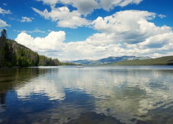 In the foreground, clouds reflect on the surface of a lake. In the center of the frame, a small boats sits in the lake. Forested hills rise on either side of the lake, and mountains, shaded by cloud cover, rise up in the distance. The sky if full of cumulus clouds, and its blue color gradually deepens in shade from the horizon upward. 