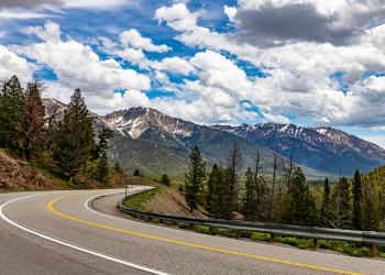 A lined, paved two-way highway winds left, right, then left again along the edge of mountains. Trees descend down from the steep incline to the left. A metal guardrail separates the road from the tree canopy rising from the downward gradient. In the background, tree covered mountains dusted with snow rise into the deep cerulean blue sky filled with puffy, white clouds.