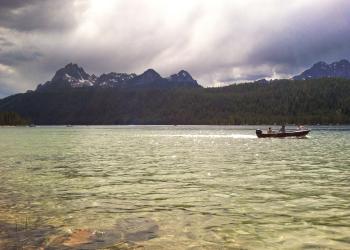 A crystal clear lake extends to the Sawtooth Mountains in the background. A small, jetted boat in the middle of the lake holds three passengers.
