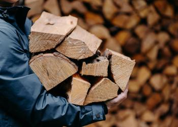 A person in a blue jacket, shown only from neck to waist, hauls seven pieces of chopped wood. In the background, a wall of chopped firewood appears out of focus.