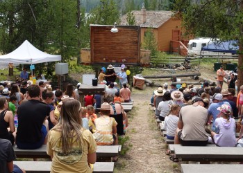 A crowd of people sit on wood benches in at the Redfish Visitor Center Amphitheatre. Smokey Bear stands in front of the crowd with a couple of ladies. Balloons pepper the background.