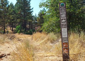 A trail sign with various recreation logos with a trail in the background.