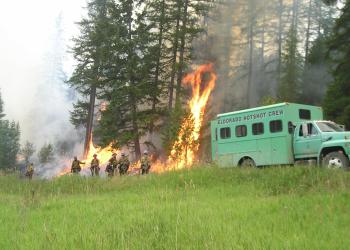 A hotshot crew of firefighters is fighting a fire along a road where their crew buggy is parked.