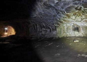 Inside of Subway Cave, a lava tube cave with smooth rock wall, dark with a flashlight shining on walls. In distance, a person surrounded by the light of their headlamp, next to the lava tube wall.