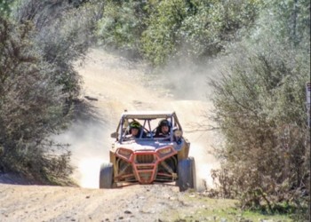 A 4-wheeled off highway vehicle with a roll cage, driving on a dirt trail surrounded by brush, oak, and shrubs. Driver and passenger both wearing helmets.