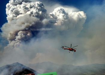 A huge cloud of smoke rises above mountains that are on fire while a large helicopter is flying by.