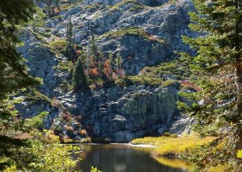 A small lake is seen beneath a rugged, rocky mountainside with trees in the foreground.