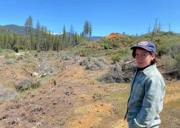 A woman stands to in the left of a photo looking towards the camera with a background of a gully with brush piled for burning and conifer trees in the background.