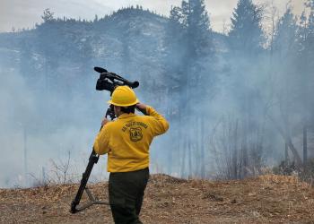a man wearing yellow nomex carries a camera on a tripod near a smokey landscape
