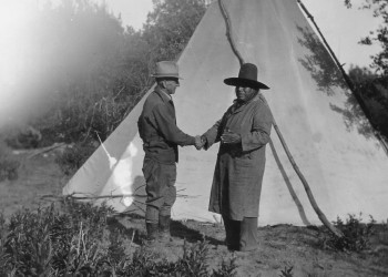 A black and white photo of two men shaking hands in front of a teepee.