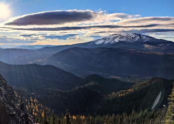 Sun shining through clouds over a valley with green and gold trees. A mountain is cloud-capped in the distance.