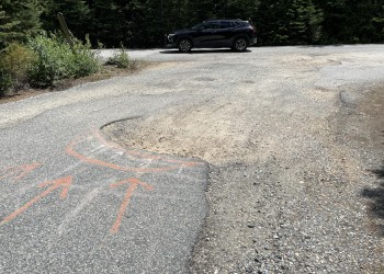 Depicted is a rough section of paved road at the entry of the trailhead parking area that has several large potholes and graveled sections of roadway.