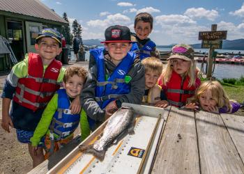 Eight kids of various edges smile at the camera from the end of a table where a fish is on a measuring device.