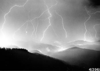 A black and white photo of lightning striking a mountainous forest.