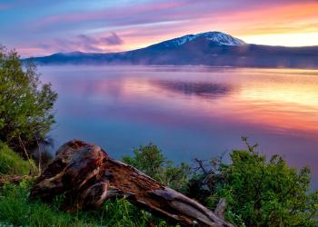 A vibrant pink and yellow sunset strikes a lake with a snow-topped mountain in the distance.