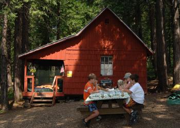 Three kids sit on at a table with snacks in front of an old, wooden building with a porch. 