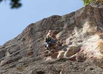 A photo of a rocky outcrop from below. Blue sky is seen in the background. A rock climber is on the rocky wall with safety gear.