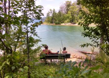 A woman and child sit fishing from a bench on the side of a lake surrounded by forest.