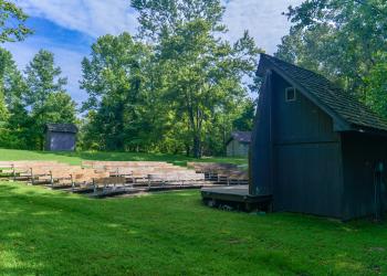A rustic amphitheater with several rows of wooden benches in front of a small stage in the forest.