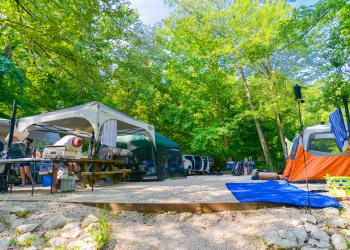 An occupied campsite in shady forest campground. A camper cooks at a table near a shade tent. Other sleeping tents and a vehicle are in the background.