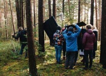 Several people holding professional filming equipment stand in a forest. 