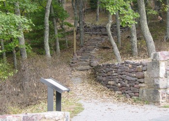 Interpretive sign with path leading past a stone wall up into a forested area. 