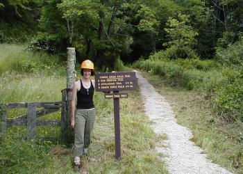 Hiker with hard hat stands next to a trail and trails sign leading to Comers Creek Falls