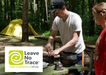 Two campers use a butane camping stove at a picnic table with the Leave No Trace logo overlayed.
