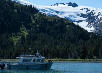 Forest Service boat anchored in front shore. Snowy and green mountains. Daylight, summer.