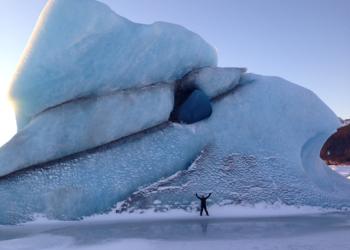 A tiny person stands with arms raised in front of a huge iceberg on a frozen lake near Sheridan Glacier