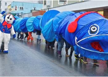A giant ice worm parade float carried by a dozen people makes its way down the street with a snowman waving from the left side of the frame.