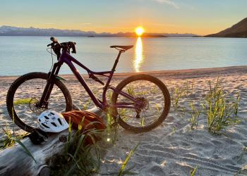 A bicycle and gear sit on a shoreline with a sunset in the background.