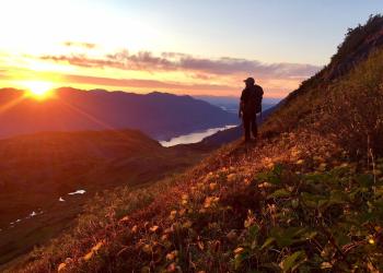A hiker pauses on a flowery hillside to view the sunset over the mountains.
