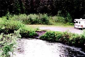 Photo of a small parking lot set by a stream. The bank of the stream is vegetated with grass and brush, except for an eroded gouge located by the parking lot.