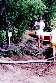 Photo of two people standing on a lakeshore with their boat. The shore is covered with dead woody debris.