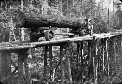 [photo] Early-day loggers transporting a freshly cut log over a wooden bridge.