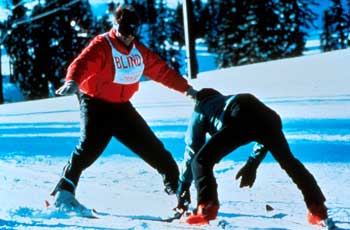 Photo of two people downhill skiing. One person is an instructor and is skiing backwards. The other person is wearing a vest that reads: BLIND.