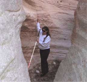 Photo of a woman with sunglasses and a cane in a slot canyon, using her free hand to search the top of the entrance to a cave in the side of the bedrock canyon wall.