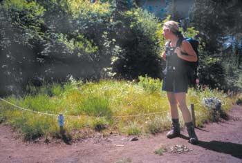 Last of three photos. This photo, taken in 1998, shows a woman standing on a trail nest to the restored campsite with a string fence. The native vegetation is lush and spreading to surrounding areas.