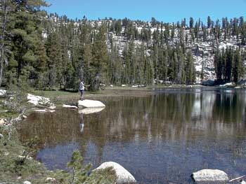 Photo of a person standing on a large rock near the shore of a mountain lake.