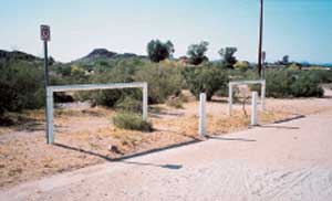 Photo of two hitch rails, near the rails are signs, bollards, and the ground around it is very uneven.