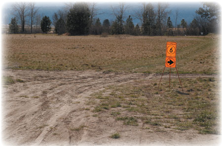 Photo of a field with tire tracks and a sign on a tripod that has a circle around a 6 and an arrow used for directions.