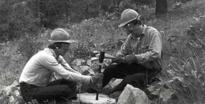 Photo of two men sitting on rocks, one is holding the drill bit on a rock and the other is hammering it.